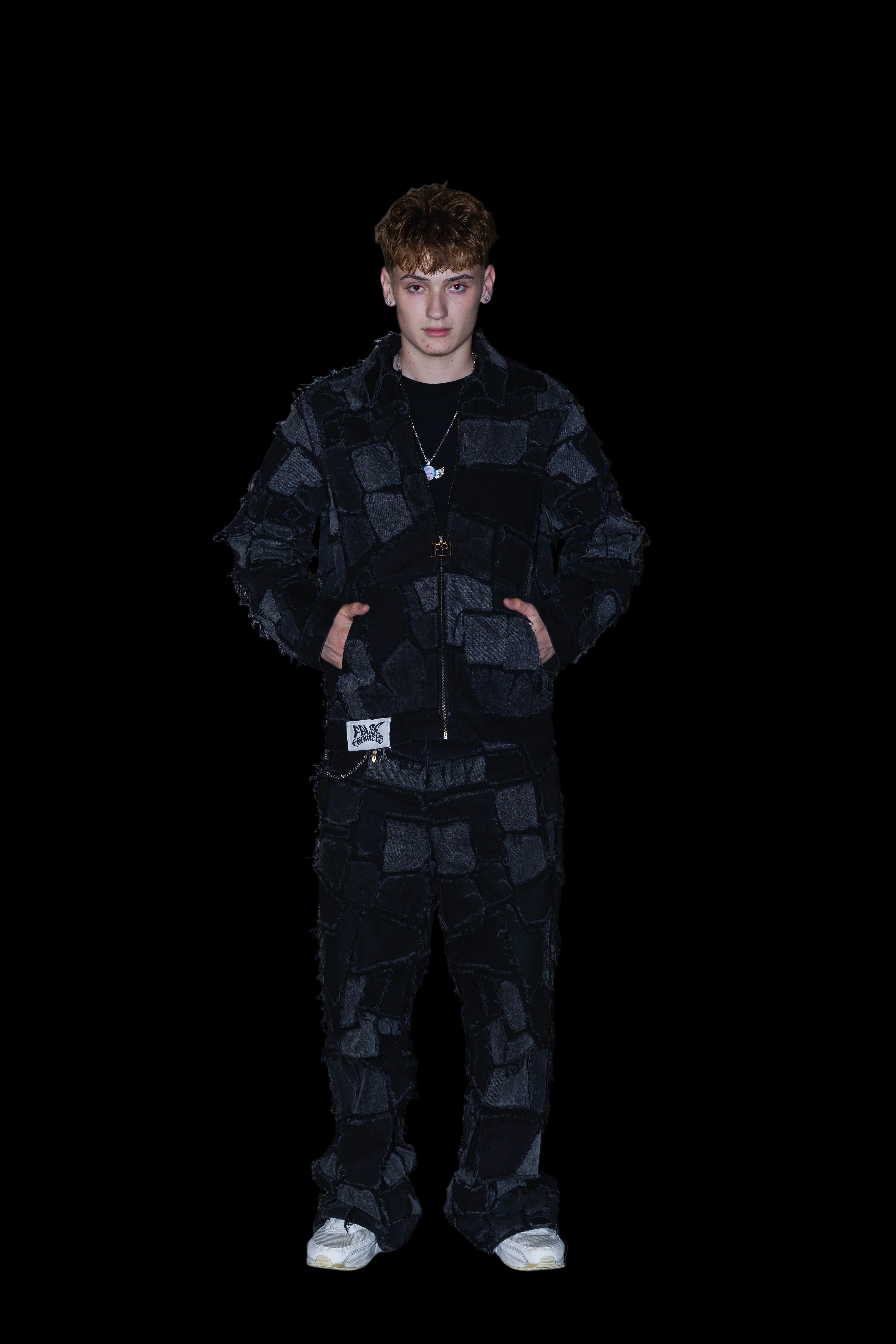 A young man wearing a black patchwork quilted jacket and matching patchwork pants, standing against a textured concrete wall.