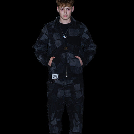 A young man wearing a black patchwork quilted jacket and matching patchwork pants, standing against a textured concrete wall.